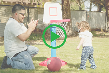 Man kneels on the grass, clapping, as his toddler plays with a basketball and a small plastic basketball hoop.