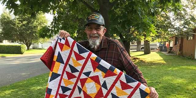 Man wearing a veteran's hat, holding up a handmade quilt and smiling