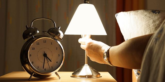 Hand turning on a lamp. A clock sits next to the lamp on a bedside table.