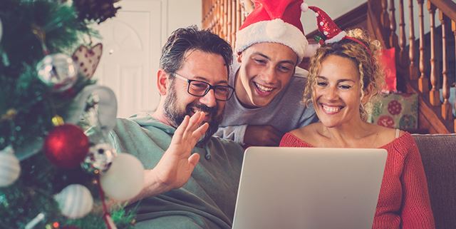 Three people are on a video call for the holidays - one man is wearing a Santa hat and the woman is wearing a headband with a smaller Santa hat. The man in glasses is waving