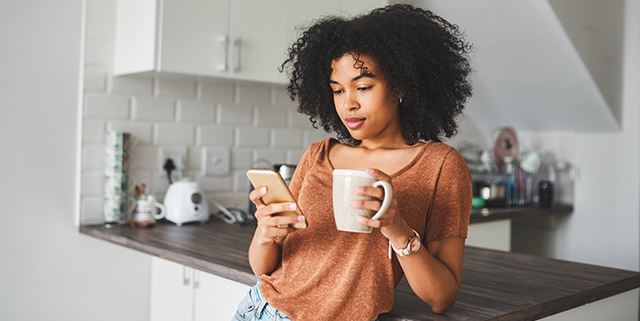 Woman on smartphone in kitchen while drinking coffee