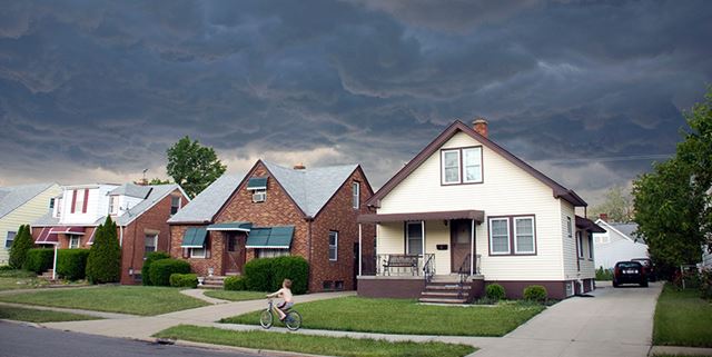 Boy rides bike on sidewalk in front of houses, rushing home as there are storm clouds in the sky