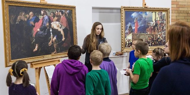 Woman standing next to collection of religious painting reproductions - children are gathered in front of them listening