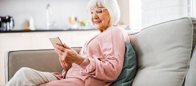 Smiling senior woman sitting on couch and looking at smartphone at home
