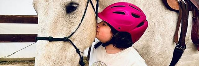 Child wearing a pink riding helmet gives a white horse a kiss on the cheek.