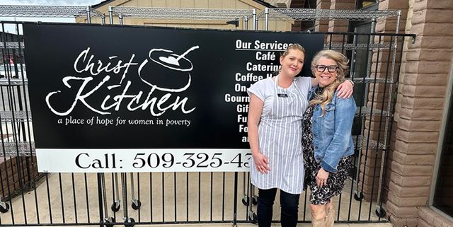Two women smiling in front of a Christ Kitchen banner. One woman is wearing an apron