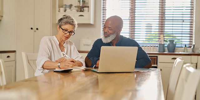 Older couple is sitting at kitchen table at home. The man is sitting in front of an open laptop and looking over at the woman, who is taking notes in a notepad