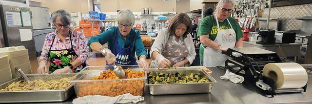 Four volunteers in a commercial kitchen preparing large meals for a meals-on-wheels program.
