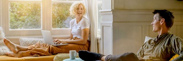 A woman is sitting on a couch next to a window at home on her laptop, she is smiling and looking towards a man who is sitting on a chair, smiling as he looks back at her.