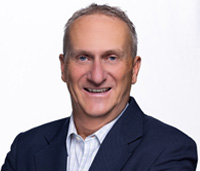 Smiling man with grey hair wearing a navy suit jacket and a white and blue striped shirt.
