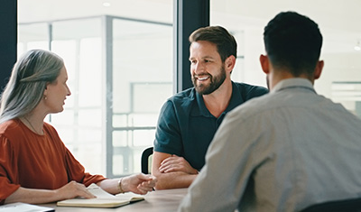 Business woman and two business men sit at a table at work, having a conversation during a team meeting.