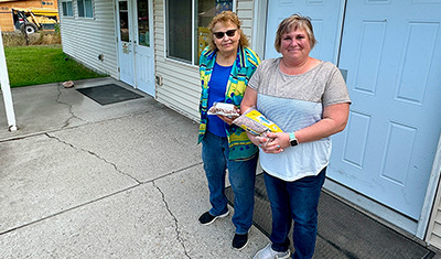 Two women stand outside on their driveway in front of their closed garage door, they are both holding bags of dried beans and rice.