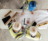 People in safety vests and hard hats look at a blueprint spread out on the table, they are pointing to it as they discuss it.