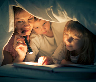 Mother, little girl, and little boy under a blanket reading a book by flashlight