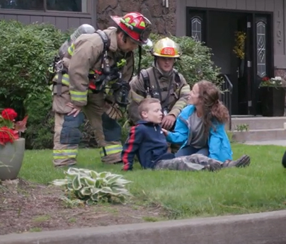 Mother and son sitting outside on grass, talking to emergency responders outside home.